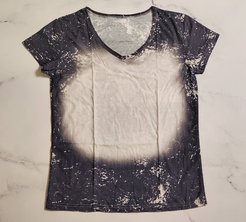 Puede incluir: Una camiseta de manga corta con cuello en V con un dise&ntilde;o &uacute;nico. La camiseta presenta un centro blanco con un patr&oacute;n gris oscuro y desgastado en los bordes. El patr&oacute;n tiene un efecto salpicado, similar a la pintura, creando una apariencia texturizada.