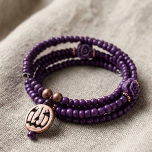 May include: A multi-strand wrap bracelet featuring small, round, deep purple beads. The design includes two purple rose-shaped beads and a round, copper-toned metal charm embossed with a jack-o'-lantern face, accented by two small copper spheres.