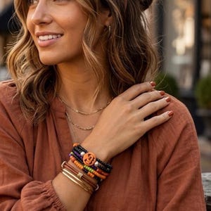 May include: A stack of bracelets featuring black, orange, and green beads, a central orange circular charm with a black letter B, and smooth gold-toned metal bangles. The jewelry is worn on the wrist alongside a rust-colored long-sleeve shirt.