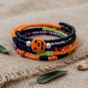 May include: A multi-strand wrap bracelet featuring small orange, black, purple, and lime green seed beads. The design includes two silver-toned metallic beads and a central round orange bead with a black carved jack-o'-lantern face.