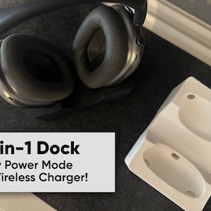 May include: A white charging dock with two indentations for headphones. The dock is on a black surface with a pair of black headphones resting on a stand. The text "2-in-1 Dock Low Power Mode + Wireless Charger!" is visible below the headphones.