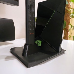 Könnte beinhalten: Ein schwarzer 3D-gedruckter Ständer mit einer schwarzen Fernbedienung und einem schwarzen Streaming-Gerät. Die Fernbedienung hat einen runden Knopf und mehrere andere Knöpfe, darunter einen Netflix-Knopf. Das Streaming-Gerät hat ein grünes Logo auf der Vorderseite.