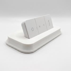 Può includere: Un telecomando rettangolare bianco con la scritta "hue" stampata, appoggiato su una base bianca abbinata. Il telecomando ha tre pulsanti con simboli relativi alla luce. La base ha un design curvo e stratificato.