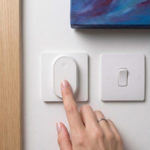 Puede incluir: Dos interruptores de luz blancos en una pared blanca. Uno tiene un botón redondeado y ovalado que está siendo presionado por un dedo. El otro es rectangular con un interruptor basculante. A la izquierda se ve un marco de madera. Los interruptores probablemente son para controlar las luces.