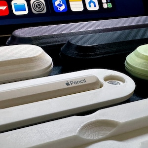 May include: A white Apple Pencil holder with the word "Pencil" printed on it. The holder is made of a smooth, white plastic and has a small, round hole on the side. The holder is sitting on a table next to other holders.