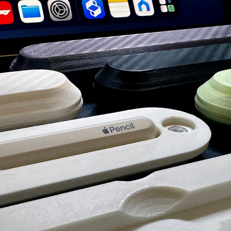 May include: A white Apple Pencil holder with the word "Pencil" printed on it. The holder is made of a smooth, white plastic and has a small, round hole on the side. The holder is sitting on a table next to other holders.
