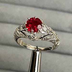 Può includere: Un anello d'argento con una grande gemma rossa rotonda. L'anello presenta un design a filigrana ornato attorno alla gemma. L'anello è tenuto da uno strumento d'argento su uno sfondo grigio.