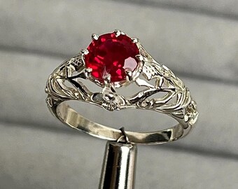 May include: A silver ring featuring a large, round, red gemstone. The ring has an intricate, filigree design. The ring is held by a silver tool against a grey background.