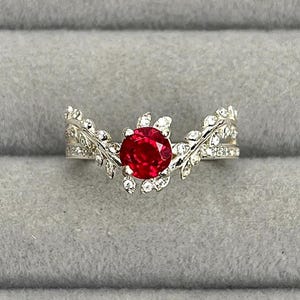 May include: A silver ring with a large, round, red gemstone at the center. The ring features a leaf-like design with small, clear gemstones. The ring is set against a gray background.