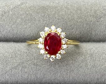 May include: A gold ring with a central oval ruby, encircled by a halo of small, round, white gemstones. The ring is displayed on a grey, textured surface.