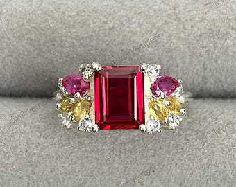 May include: A silver ring with a striking design. It features a large, rectangular, deep red gemstone at its centre, complemented by smaller, round, pink gemstones and clusters of yellow and white stones, creating a vibrant and elegant look.