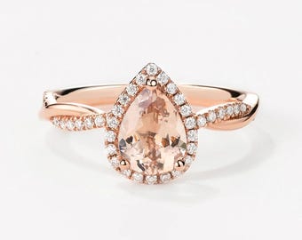 May include: A rose gold engagement ring with a pear-shaped morganite center stone and a halo of smaller diamonds. The band is twisted and features a row of smaller diamonds.