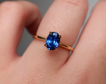Puede incluir: Un anillo de oro con una vibrante gema azul ovalada. La gema está sujeta por cuatro garras. La banda del anillo es de oro fino y pulido. El anillo es una pieza de joyería.
