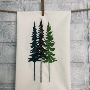 May include: White kitchen towel with an embroidered design of three green, blue, and black pine trees.