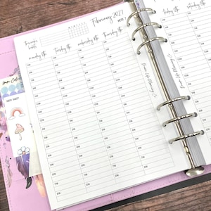 May include: A ring-bound planner open to a February 2027 weekly schedule page with hourly time slots from 5 AM to 8 PM. The planner features a pink cover, a deco sticker sheet with a rainbow and mushroom, and the text Dream Life Planning Co.
