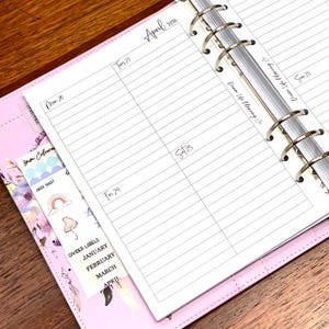 May include: A planner open at April 2026, showing lined pages for daily scheduling. A decorative bookmark with stickers and divider labels is visible. The planner has a pink cover and silver rings.