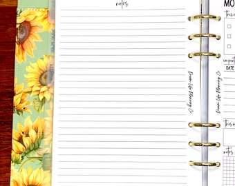 May include: A white planner with a gold ring binding. The planner has a notes section with lined paper and a monthly calendar section. The planner is decorated with a sunflower pattern.