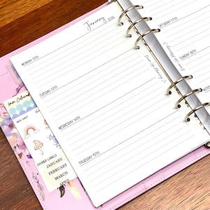 May include: A pink planner open to a January 2026 weekly spread. The pages are lined with dates for each day of the week. A pocket holds decorative stickers and divider labels. The planner is on a wooden surface.