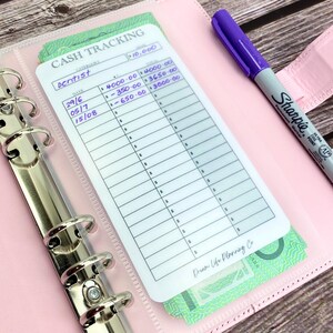May include: A white cash tracking sheet with a purple pen and a stack of Australian dollar bills. The sheet has a column for date, category, and amount. The category is "Dentist" and the amount is £10,000. The sheet is labelled "Cash Tracking".