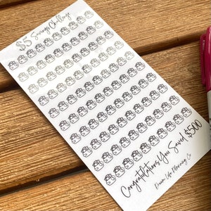 May include: A sheet of stickers featuring a black and white illustration of a piggy bank. The stickers are arranged in rows and columns. The text "$5 Savings Challenge" and "Congratulations You Saved £500" is printed on the sheet. The text "Dream Life Planning Co" is printed in the bottom right corner.