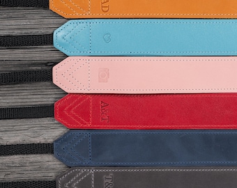 May include: A collection of personalized leather camera straps in various colors, including brown, orange, blue, pink, red, navy, gray, and black. Each strap features initials or a word, such as "DAD", and decorative stitching.