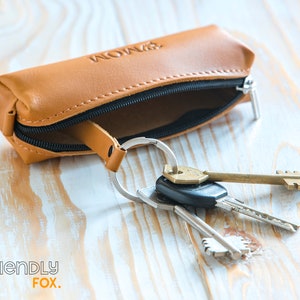 May include: A tan leather key case with a black zipper and the word "MOM" embossed on the front. A silver keyring holds several keys of different shapes and sizes. The case is open, revealing the interior. The background is a light-colored wooden surface.