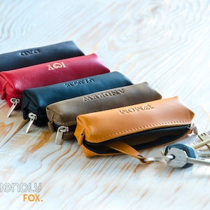 May include: Five leather keychains with zipper closures in different colors, each with a personalized name engraved on the front. The keychains are lined up on a light wood surface. The keychain at the front has a set of keys attached.