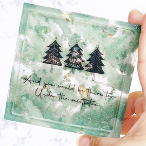 May include: A green and gold glitter resin coaster with three black silhouette Christmas trees and the text "And you would be there too Under the mistletoe".