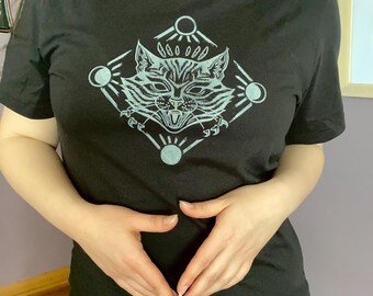 May include: Black t-shirt featuring a detailed, light blue graphic of a cat's face within a diamond shape, surrounded by sun and moon symbols. The cat has an open mouth and sharp claws. The shirt has short sleeves.