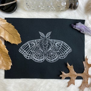 May include: A black fabric patch with a white embroidered moth design. The moth has large wings with intricate details and a fuzzy body.