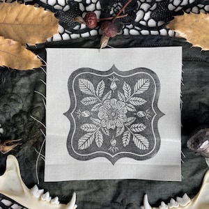 Peut inclure: Un carré de tissu blanc avec un motif floral noir imprimé dessus. Le motif représente une rose stylisée avec des feuilles et des boutons. Le tissu est texturé et a un léger brillant.
