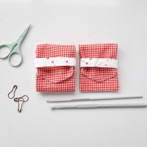 May include: Two red and white gingham fabric pouches with white fabric flaps and a row of colourful pins. The pouches are sitting on a white surface next to a pair of teal scissors, three safety pins, and two grey knitting needles.