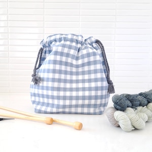 May include: A blue and white gingham drawstring bag with grey rope ties and tassels. Wooden knitting needles and several skeins of yarn in shades of blue and grey are next to the bag. The bag is for storing knitting supplies.