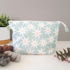 May include: A zipped pouch featuring a light green fabric with a repeating pattern of white daisy-like flowers with yellow centres. The pouch has a white zip closure and a small fabric tab on the side, showcasing a clean and functional design.