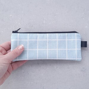 May include: A light blue pencil case with a white grid pattern and a black zipper. The case has a black strap.