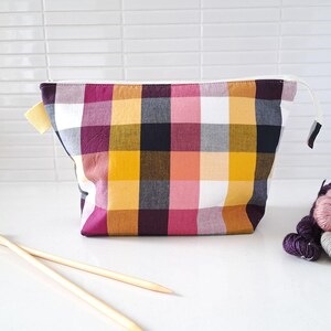 May include: A zipped pouch featuring a multi-coloured plaid pattern in shades of mustard yellow, deep magenta, black, and white. The fabric bag has a rectangular shape with a white zip closure and a small yellow fabric tab on the left side.
