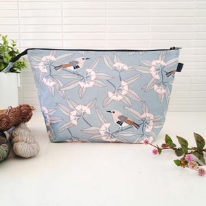 May include: A trapezoidal zipped pouch featuring a light blue fabric with a repeating pattern of small birds perched on branches with pale pink flowers and grey leaves. The pouch has a black zip closure along the top edge.