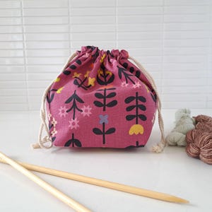 May include: A drawstring pouch in a deep mauve fabric featuring a repeating pattern of stylised black stems with yellow, pink, and light blue flowers. The bag is cinched with thick cream-coloured rope cords and sits near two wooden knitting needles.