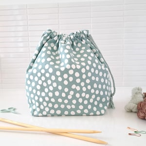 May include: A sage green drawstring project bag featuring a white polka dot pattern. The bag is cinched at the top with a matching sage green braided cord. The fabric has a soft texture and the bag stands upright with a rounded base structure.