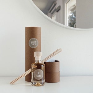 Könnte beinhalten: Eine braune Pappröhre mit einem weißen Etikett, auf dem "Luce dolce" steht, und eine klare Glasflasche mit einem Korkverschluss und einem weißen Etikett, auf dem "Be Happy" steht. Die Flasche ist mit einer hellbraunen Flüssigkeit gefüllt und im Hintergrund befinden sich Holzstäbchen.