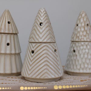 Könnte beinhalten: Drei keramische Weihnachtsbaum-förmige Wachs-Wärmer. Die Bäume sind weiß mit verschiedenen Mustern. Der Baum links hat ein gestreiftes Muster, der mittlere Baum ein Zickzackmuster und der Baum rechts ein strukturiertes Muster.