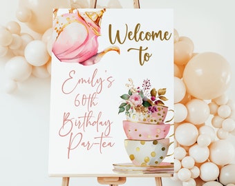 May include: Welcome to Emily's 60th Birthday Par-tea sign with a pink teapot and stacked teacups with gold accents and floral decorations.