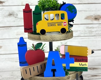 May include: A two-tiered wooden tray with a black metal stand. The top tier has a yellow school bus, a green crayon, a red crayon, a globe, and a potted plant. The bottom tier has a blue crayon, a red apple, a ruler, a blue letter A, a yellow pencil, and a stack of books that say "Reading", "Math", and "Science".