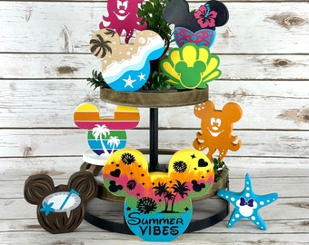 May include: A tiered wooden tray with a variety of painted wooden Mickey Mouse ears. The ears are decorated with summer themes, including palm trees, seashells, and a starfish. The tray also features a sign that reads "Summer Vibes".