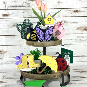 May include: A two-tiered wooden tray with garden-themed decorations. The top tier features a bee, butterfly, birdhouse, and pink flowers with tulips. The bottom tier has a ladybug, umbrella, boots, and a wheelbarrow. A shovel is also included.