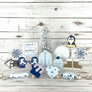 May include: A collection of winter-themed wooden ornaments, including a penguin, a snowman, mittens, a snowflake, a popsicle, a heart, a ski mask, a hat, boots, and a sign that reads "winter kisses snowflake wishes".