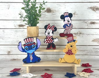 Può includere: Espositore in legno con sagome di personaggi dei cartoni animati di Topolino, Minnie, Stitch e Winnie the Pooh, tutti in abiti patriottici. Accenti decorativi a forma di stella in rosso, bianco e blu sono sparsi sotto.