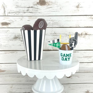 May include: A white cake stand with a scalloped edge holds a black and white striped paper cone filled with two brown football-shaped treats and a small green metal bucket with the words "Game Day" and a football on top.