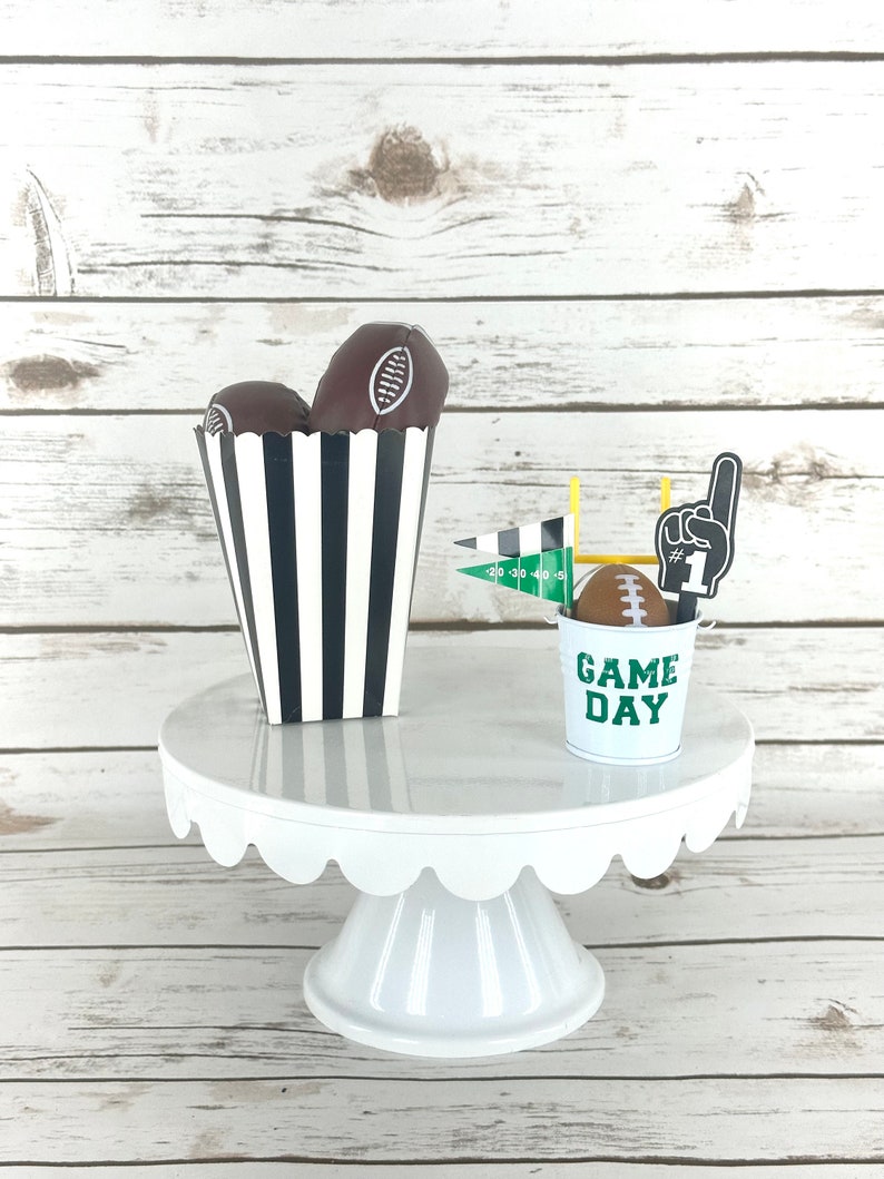 May include: A white cake stand with a scalloped edge holds a black and white striped paper cone filled with two brown football-shaped treats and a small green metal bucket with the words "Game Day" and a football on top.