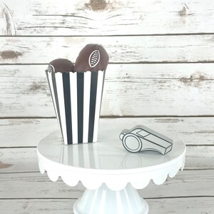 May include: A white cake stand with a scalloped edge, decorated with a black and white striped box filled with chocolate footballs and a silver whistle.